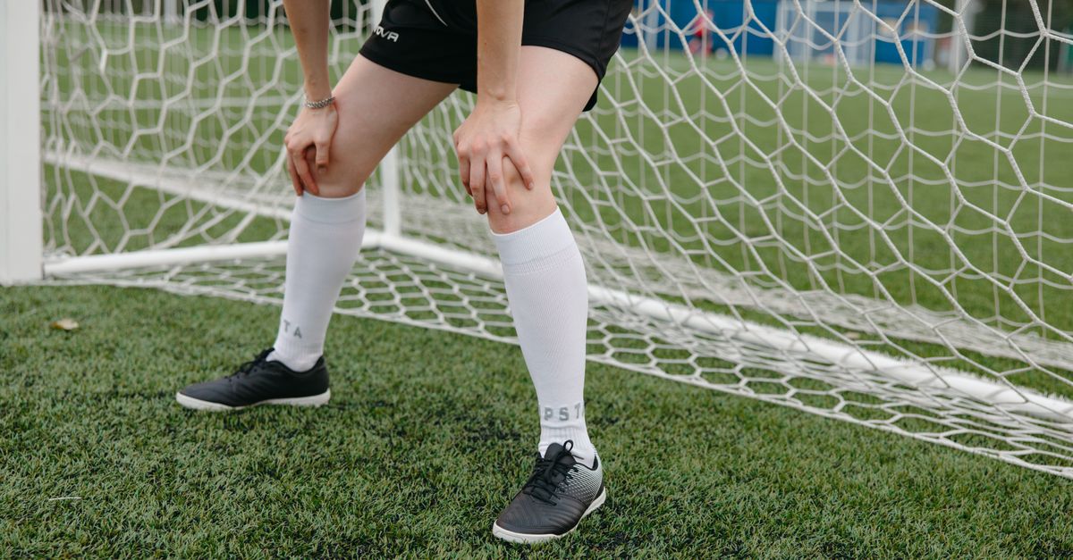 Soccer player resting in front of the goal on a grassy field, wearing sports uniform and sneakers.